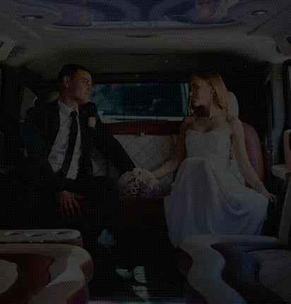 A bride and groom pose together beside a white bus, smiling and dressed in wedding attire.